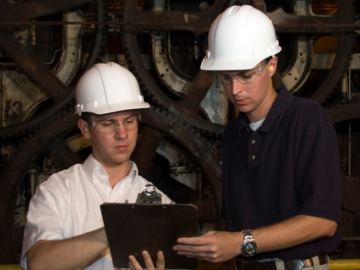 Two workers in hard hats reviewing safety checklists on clipboards, ensuring machine guarding compliance and workplace safety in an industrial setting.