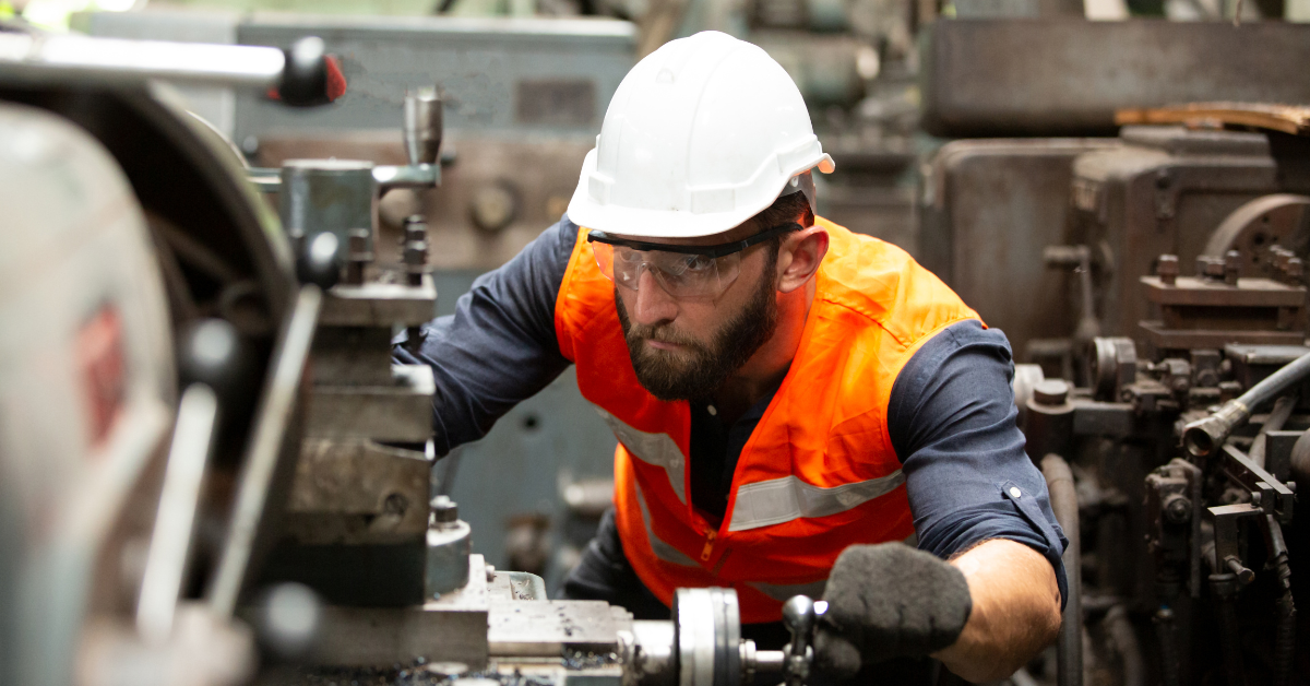 Worker in a machine shop wearing a safety hat, glasses, and vest, ensuring compliance with workplace safety regulations while operating industrial equipment.